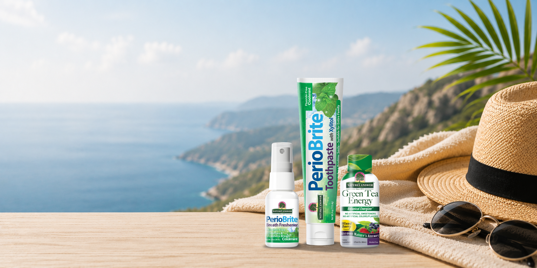 PerioBrite oral care products are displayed on a wooden table in front of a scenic coastal background. The products include a toothpaste tube, a breath freshener spray bottle, and a green tea energy bottle, styled with a straw hat, sunglasses, and a towel alongside the beach view.