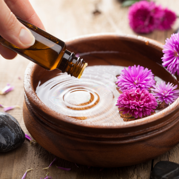 A hand carefully pouring essential oil into a wooden bowl filled with water and floating purple flowers, set on a natural background.